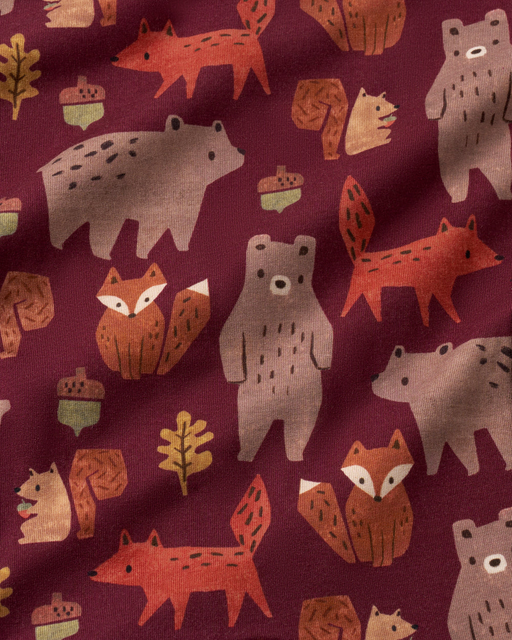 A close-up of a fabric surface with an all-over autumn woodland print featuring stylized foxes, bears, squirrels, acorns, and leaves on a deep burgundy background.