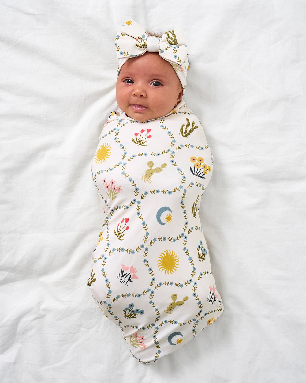 Baby on a white bed wearing the Desert Meadow Swaddle & Luxe Bow Headband Set