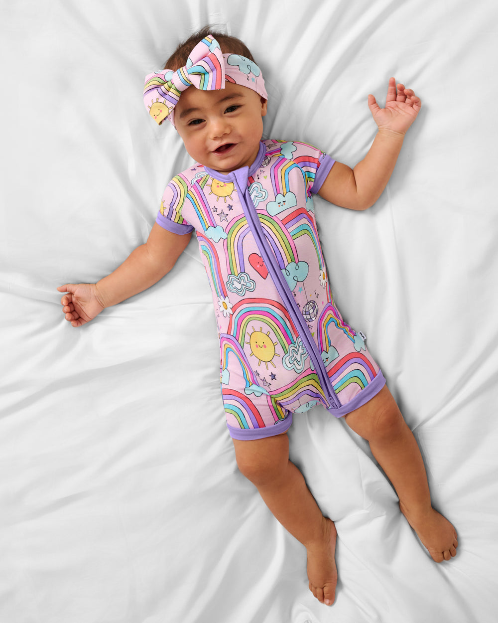 Aerial view of baby wearing the Disco Rainbows Shorty Zippy with a matching luxe bow headband