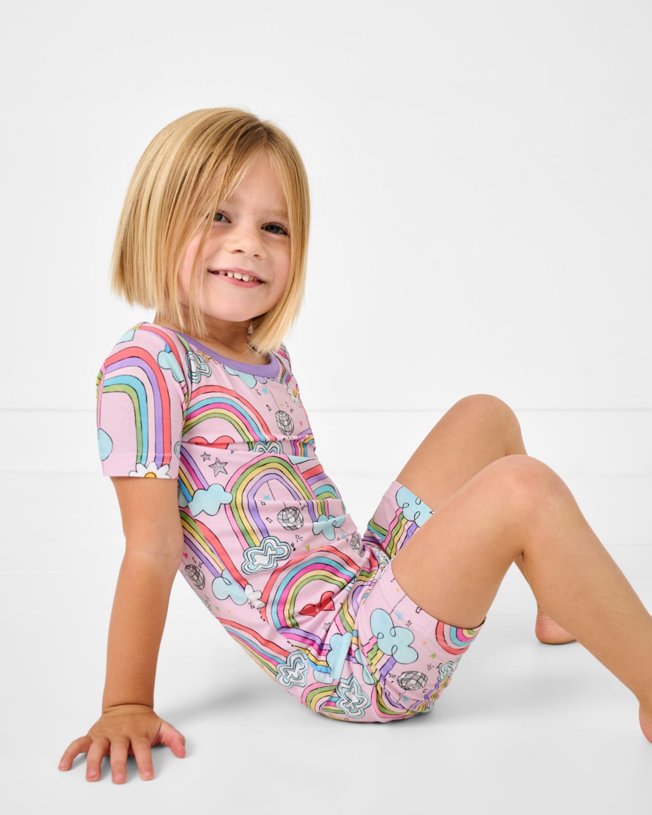 Sitting girl wearing the Disco Rainbows Two-Piece Short Sleeve & Shorts Pajama Set
