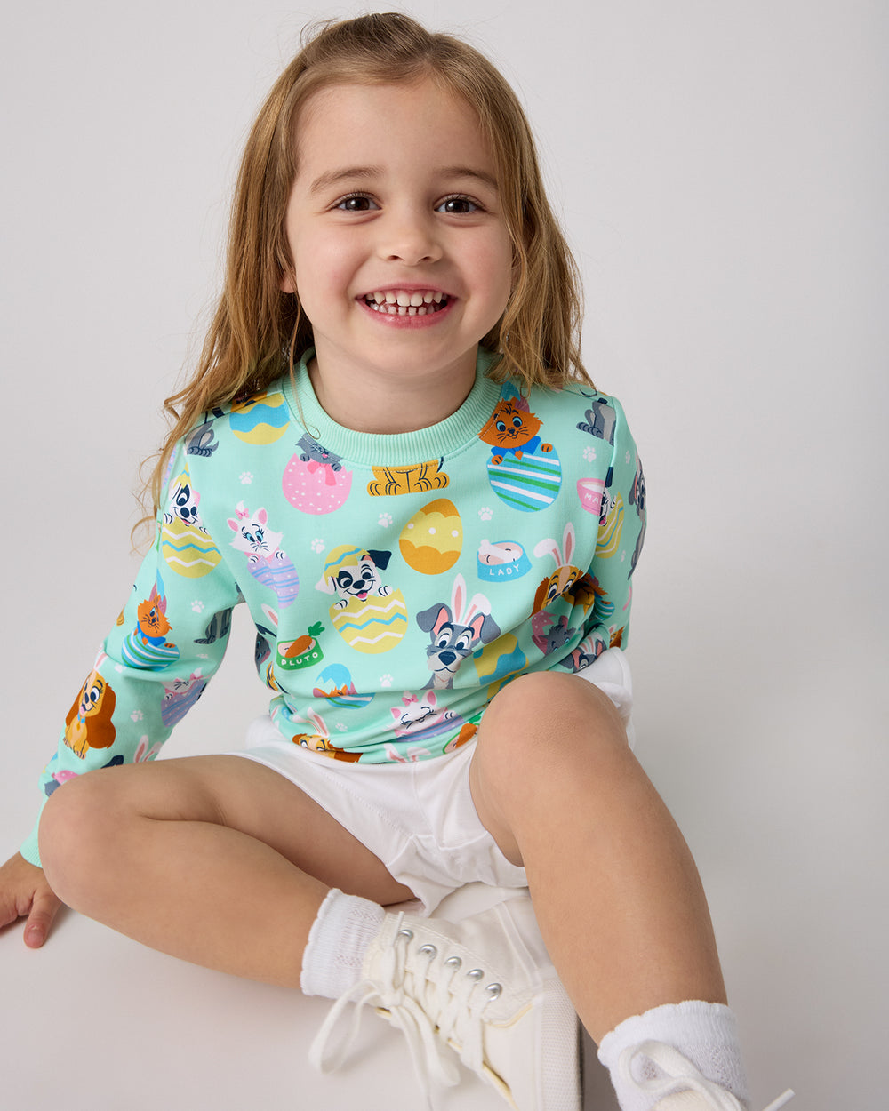 Sitting girl wearing the Disney Peekaboo Pets Crewneck Sweatshirt with white denim shorts