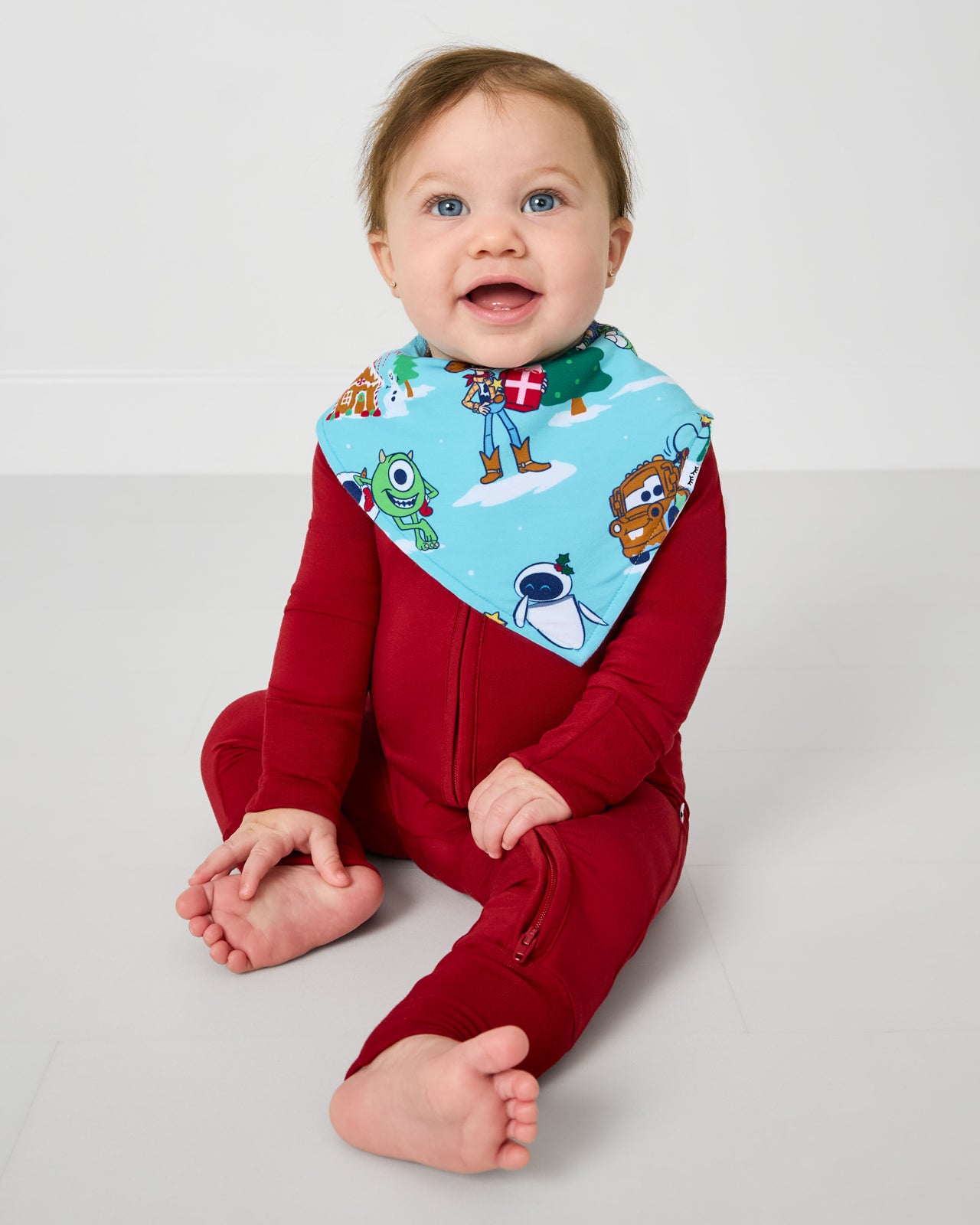 Sitting baby wearing the Disney & Pixar Christmas Village Bandana Bib with a red zipper pajama