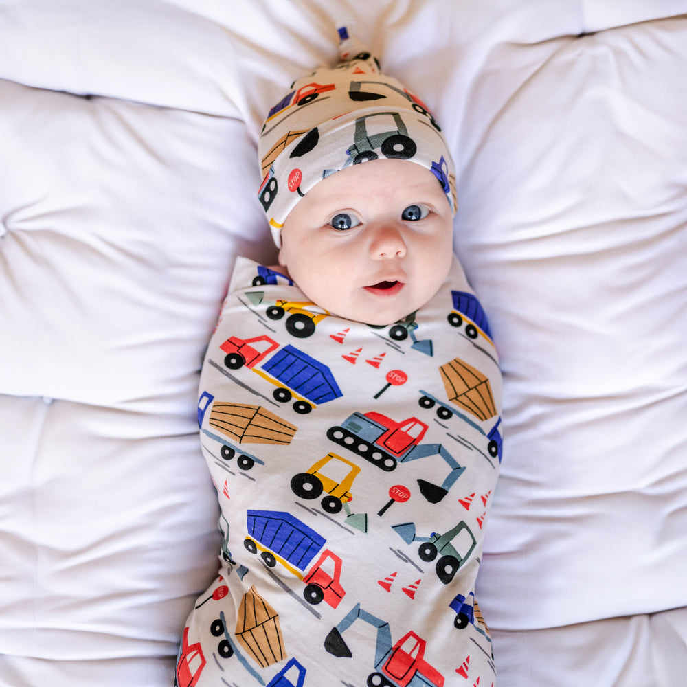 Click to see full screen - Image of infant baby laying in a bassinet basket. He is shown wearing a construction printed swaddle and hat set. This print features utility trucks and tractors on a white background.