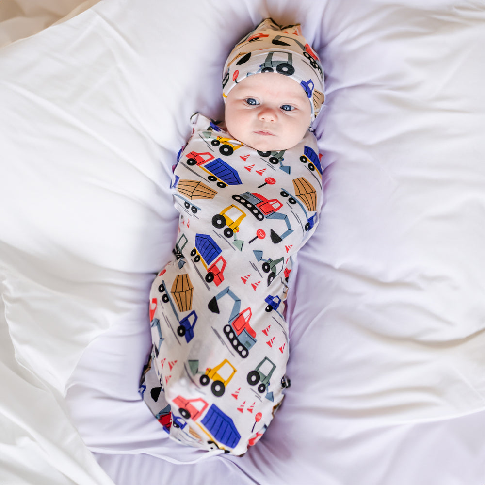 Click to see full screen - Image of infant baby laying in a bassinet basket. He is shown wearing a construction printed swaddle and hat set. This print features utility trucks and tractors on a white background.