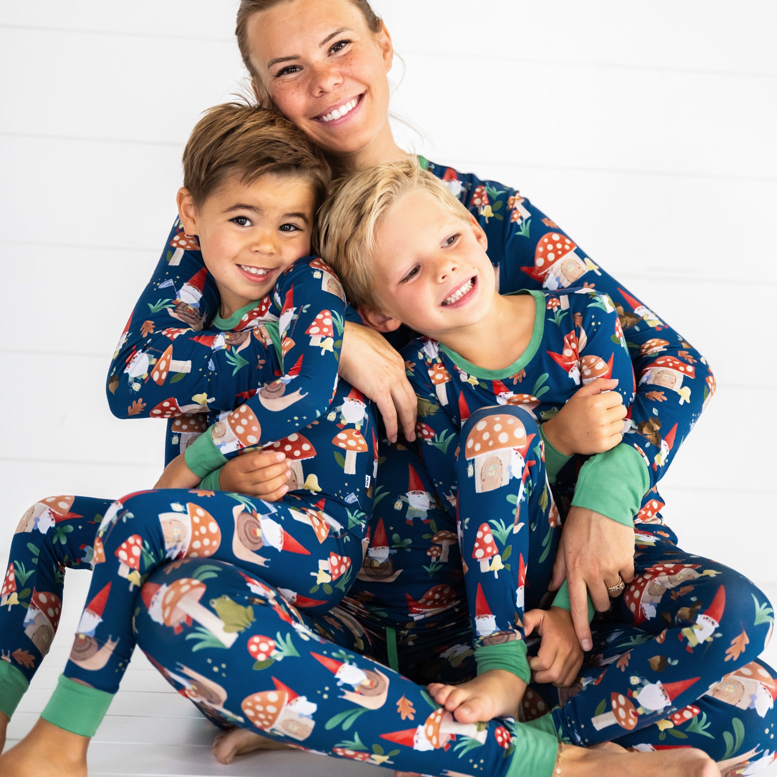 Smiling family of three wearing the Forest Gnomes print. Female model is wearing the Forest Gnomes Women's Pajama Pant and Top while hugging two boys wearing the Forest Gnomes Two-piece Pajama Set