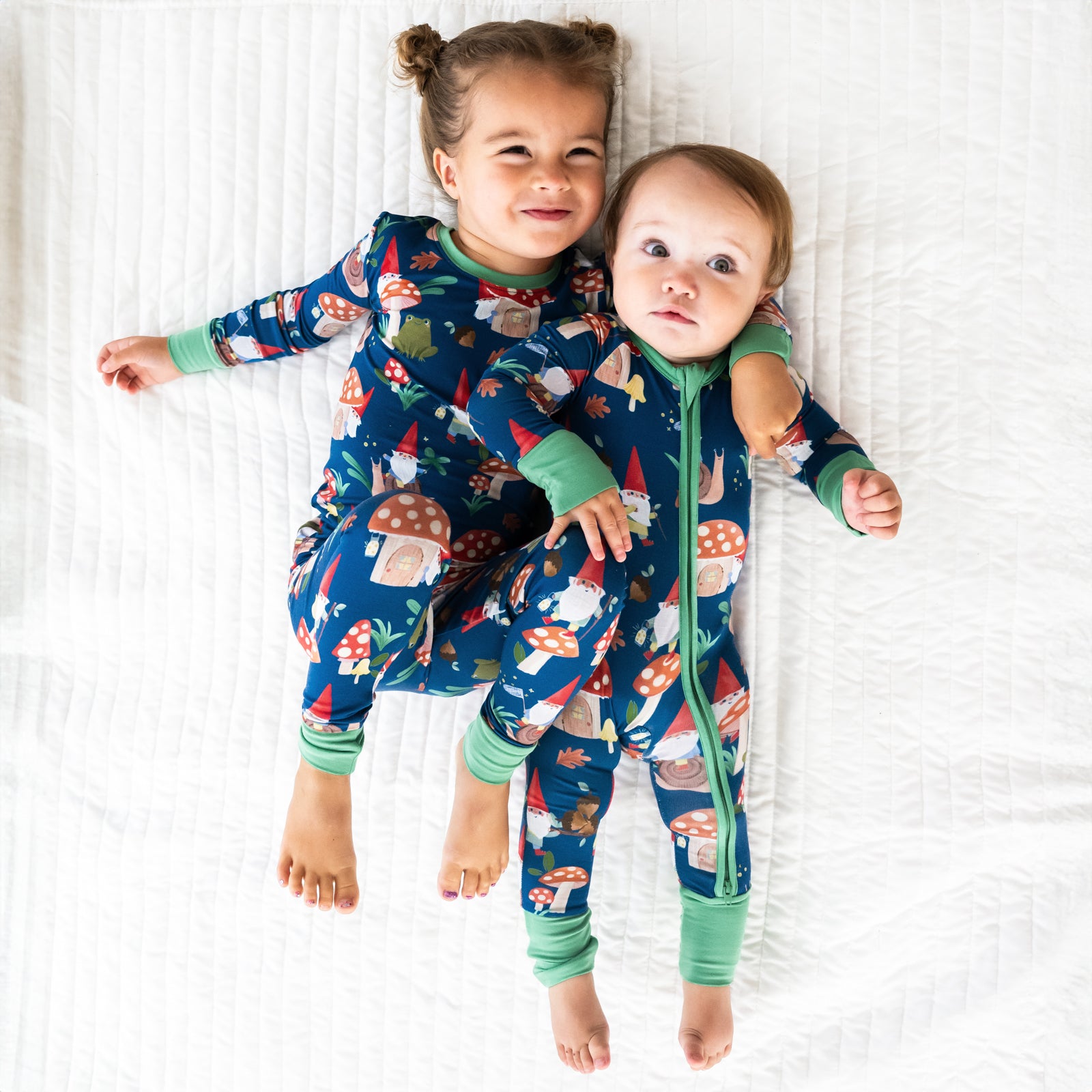 Top view image of two children laying down while wearing the Forest Gnomes print. Girl on the left is wearing the Forest Gnomes Two-piece Pajama Set and baby on the right is in the Forest Gnomes Zippy