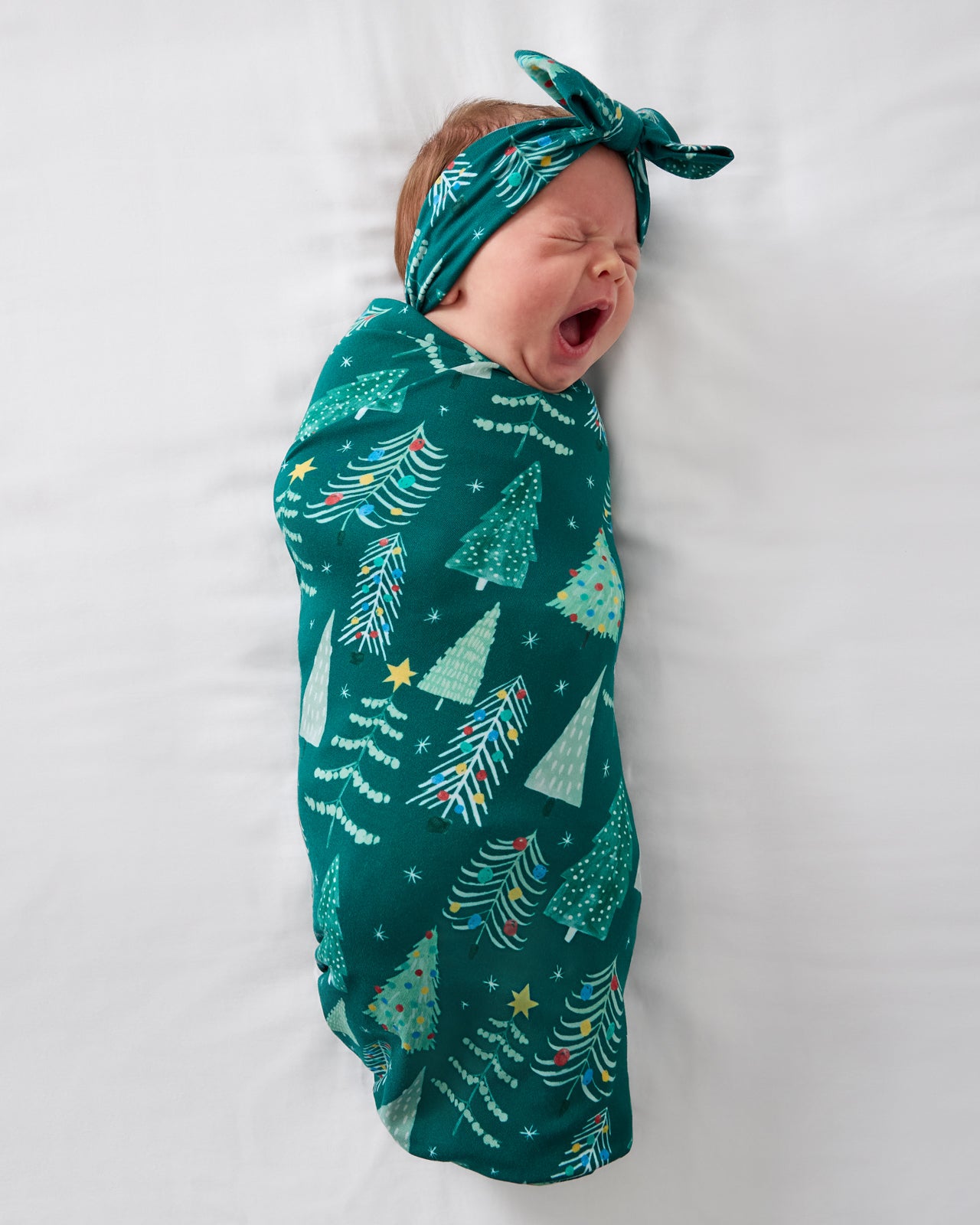Aerial view of baby wearing the Twinkling Trees Swaddle & Luxe Bow Headband Set