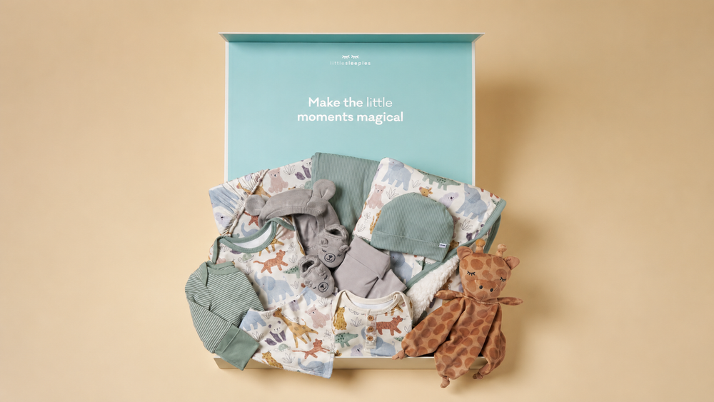 A curated baby gift set arranged in an open box against a warm beige background. The set includes a light blue printed baby onesie, a gray plush toy, a gray hat, a green fabric pouch, and a brown giraffe plush toy.
