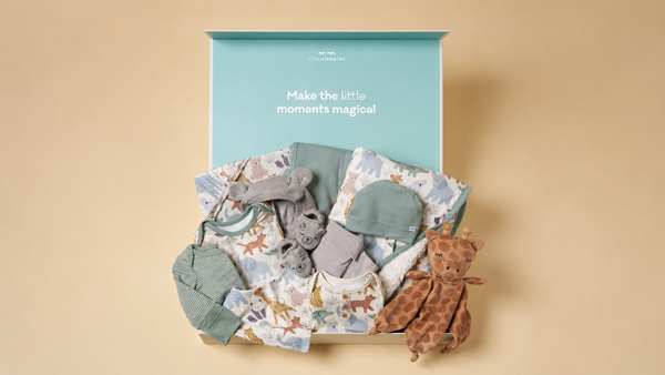 A curated baby gift set arranged in an open box against a warm beige background. The set includes a light blue printed baby onesie, a gray plush toy, a gray hat, a green fabric pouch, and a brown giraffe plush toy.