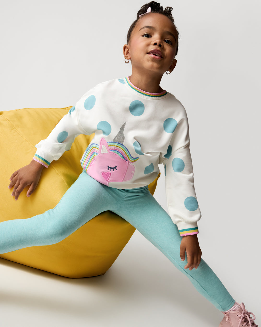 Girl wearing styles from the Winter Play drop on a yellow ottoman