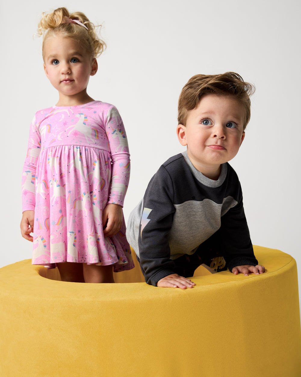 Two children wearing styles from the Winter Play collection on a yellow ottoman