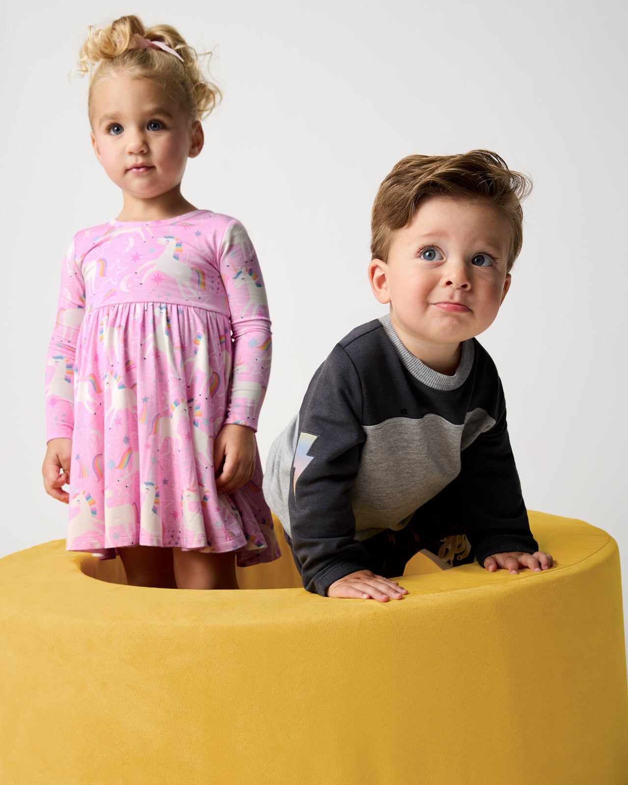Two children wearing styles from the Winter Play collection on a yellow ottoman