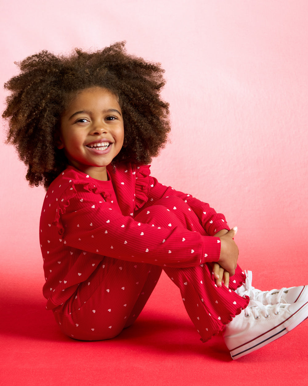 Sitting girl wearing the Little Love Ribbed Flare Pant and Crewneck on a red background