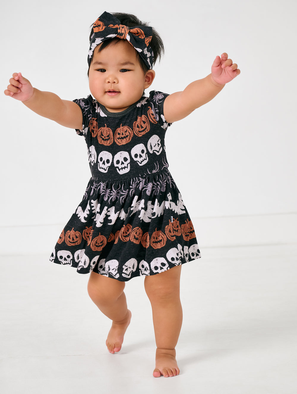 Walking toddler wearing the Hallows Eve Skater Dress with Bodysuit with a matching bow headband