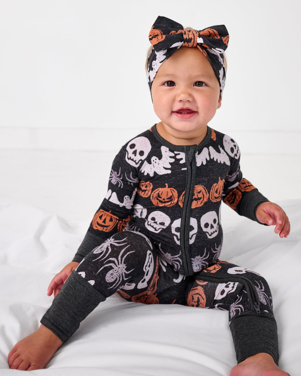 Siting baby wearing the Hallows Eve Luxe Bow Headband with a matching zippy