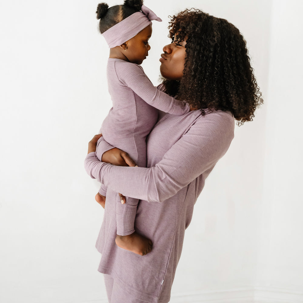 Alternate view of a mother holding up her child. Mom is wearing a women's Heather Smokey Lavender Ribbed pajama top and matching women's pajama pants. Her baby is matching wearing a Heather Smokey Lavender ribbed crescent zippy and luxe bow headband