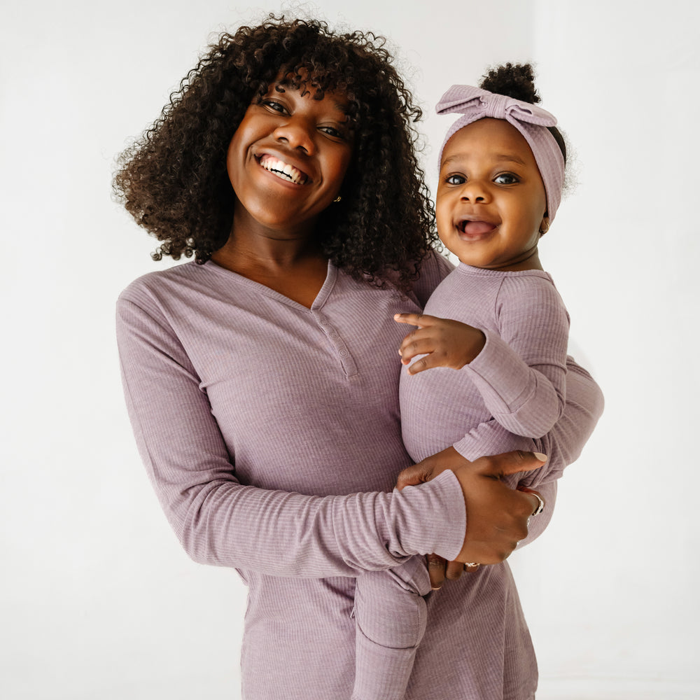 Click to see full screen - Mother holding up her child. Mom is wearing a women's Heather Smokey Lavender Ribbed pajama top and matching women's pajama pants. Her baby is matching wearing a Heather Smokey Lavender ribbed crescent zippy and luxe bow headband