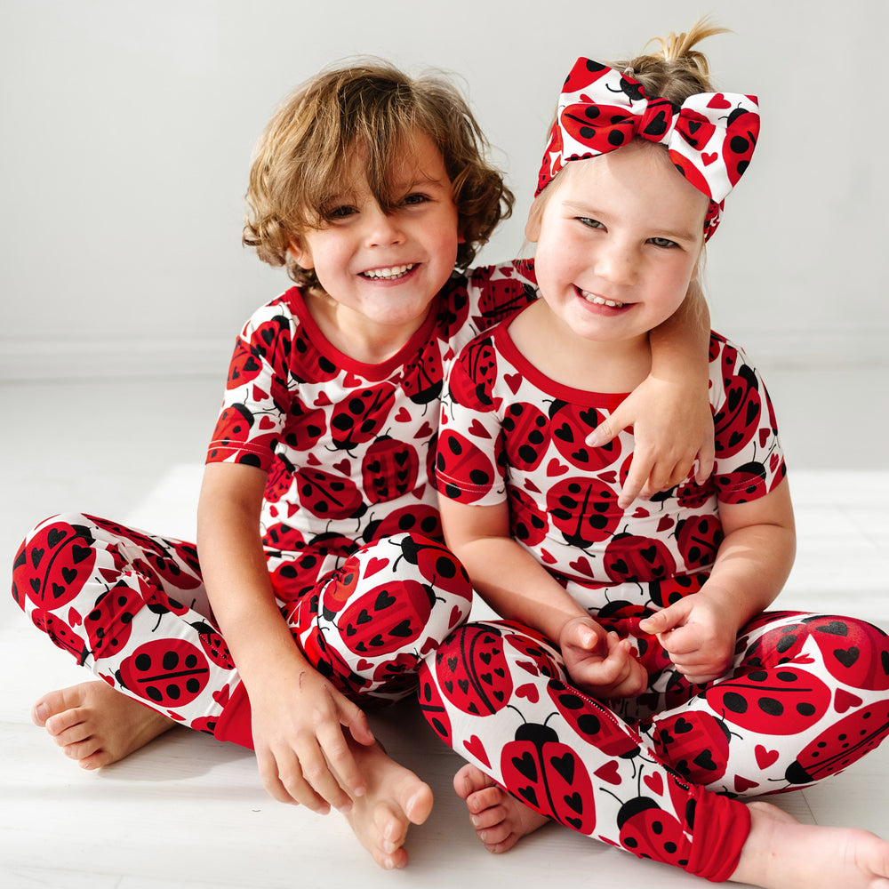Click to see full screen - Two children sitting on the ground, one wearing a Love Bug printed luxe bow headband and both wearing matching Love Bug pajamas