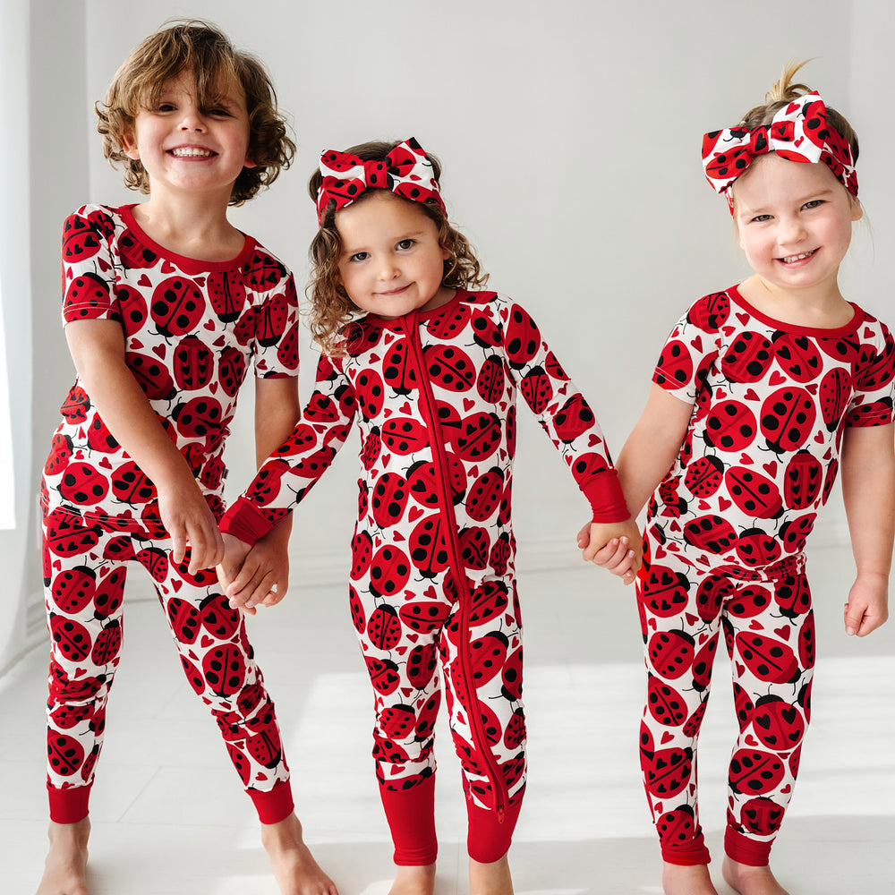Click to see full screen - Three children holding hands, two are wearing Love Bug printed luxe bow headbands and all wearing matching Love Bug pajamas