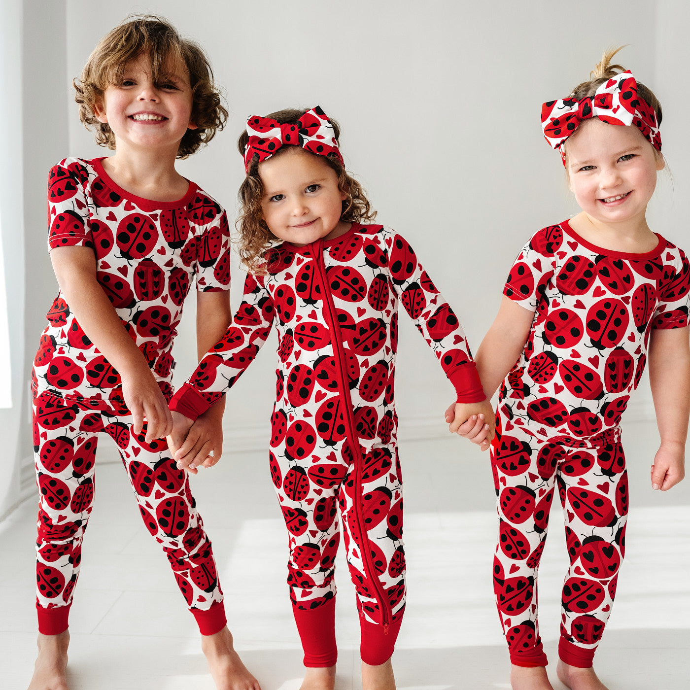 Three children holding hands, two are wearing Love Bug printed luxe bow headbands and all wearing matching Love Bug pajamas