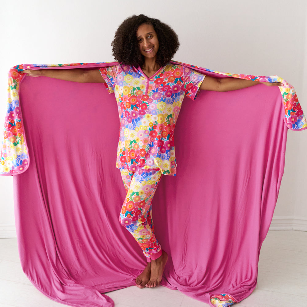 Woman holding out a Rainbow Blooms oversized cloud blanket behind her, detailing the solid pink backing and wearing matching pajamas