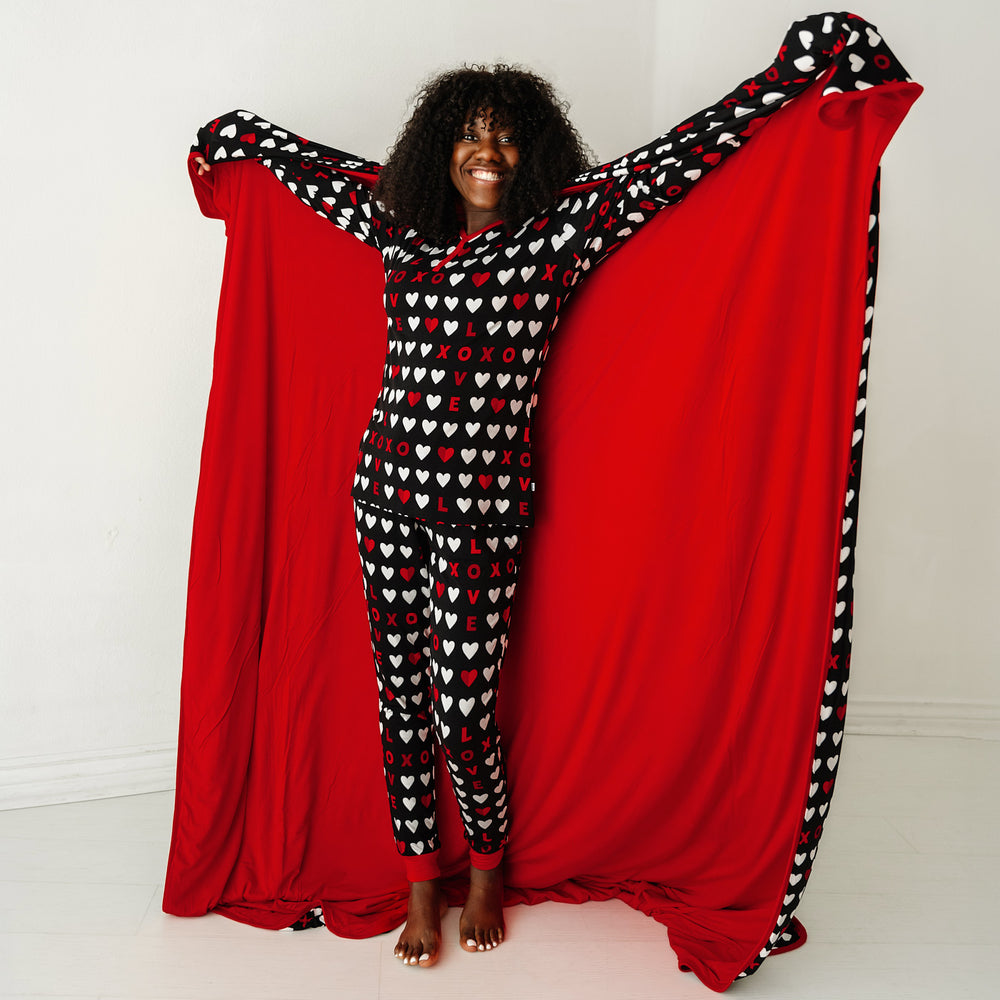 Click to see full screen - Image of a woman holding up a Black XOXO Oversized Cloud Blanket showing off the solid red backing. She is wearing women's Black XOXO pajama top and matching pajama pants