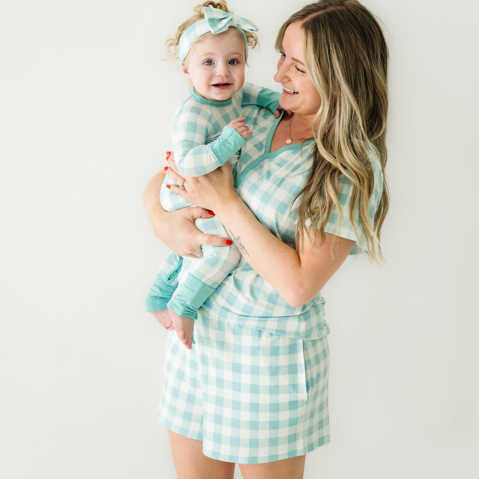 Woman holding her child wearing matching Pink Gingham pajamas. Mom is wearing women's Aqua Gingham pajama shorts and matching women's short sleeve pajama top. Child is wearing a matching Aqua Gingham crescent zippy paired with a matching luxe bow headband