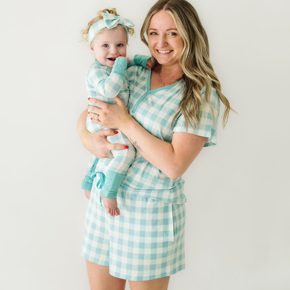 Click to see full screen - Woman holding her child wearing matching Aqua Gingham pajamas. Mom is wearing women's Aqua Gingham pajama shorts and matching women's short sleeve pajama top. Child is wearing a matching Aqua Gingham zippy paired with a matching luxe bow headband