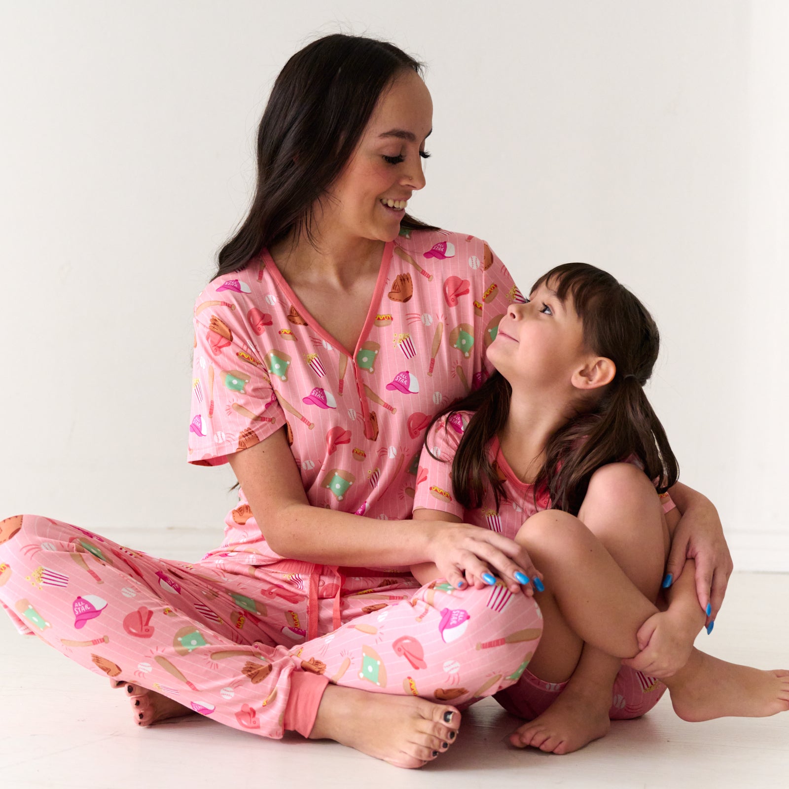 Mother and child sitting together wearing matching Pink All Stars pajamas. Mom is wearing women's Pink All Stars pajama top an matching pajama pants. Child is matching wearing a two Pink All Stars two piece short sleeve and shorts pajama set