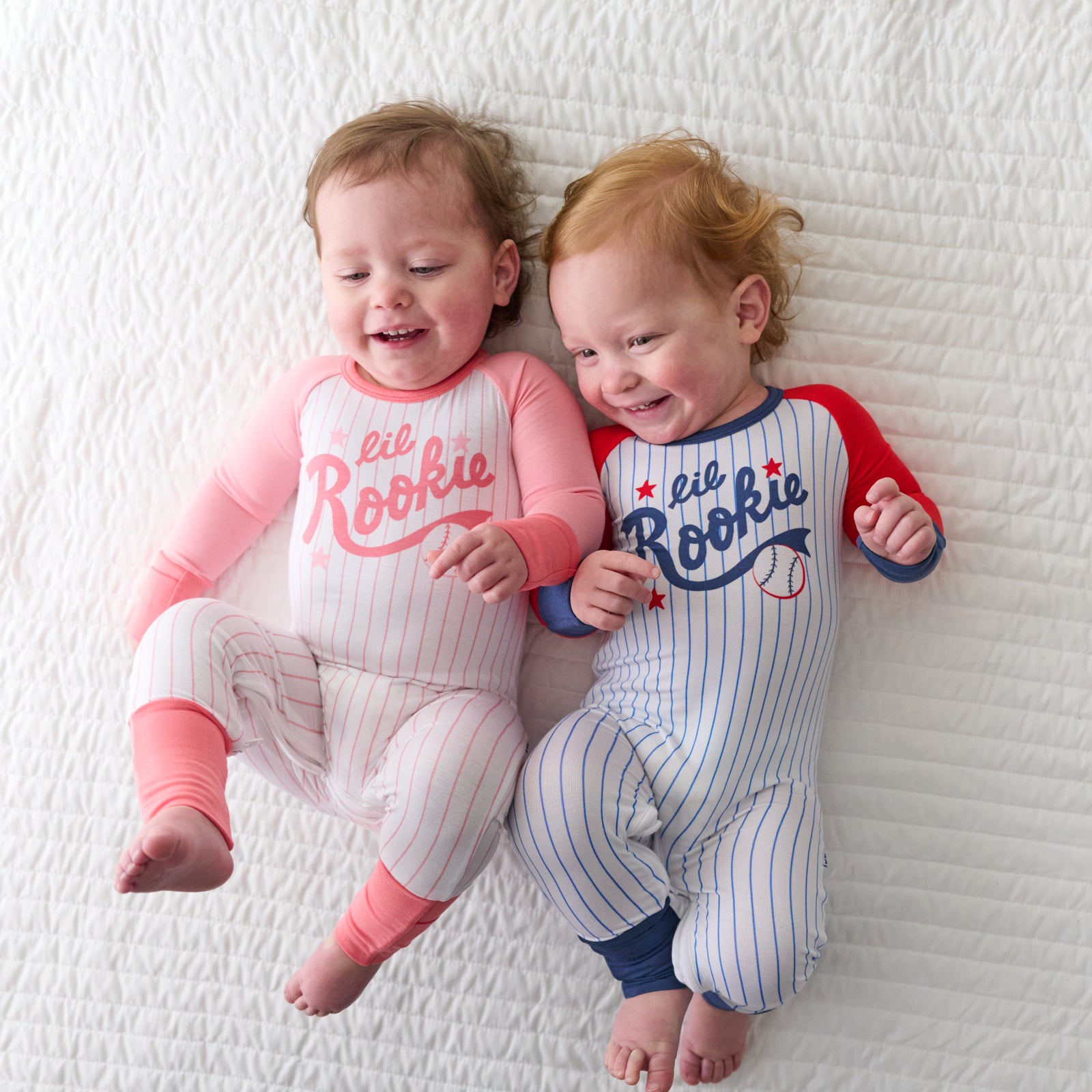 Two babies laying on a bed wearing coordinating pink and blue Lil Rookie crescent zippies