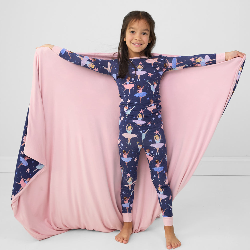 Image of a child holding out a Twilight Ballet cloud blanket showing the solid pink backing. She is wearing a matching two piece pj set