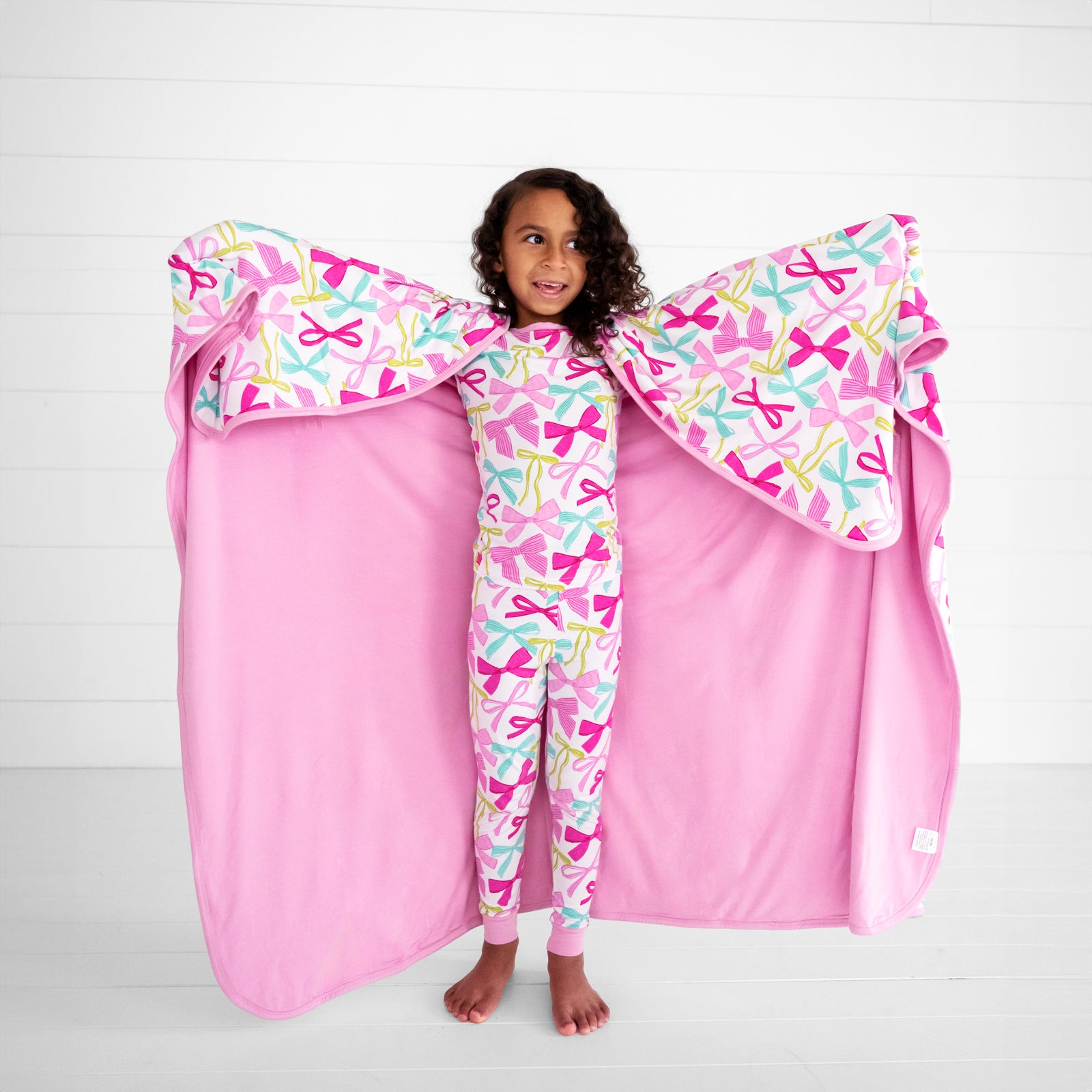 Child wearing a Ribbons and Bows two piece pj set holding out a matching luxe cloud blanket showing the solid pink backing