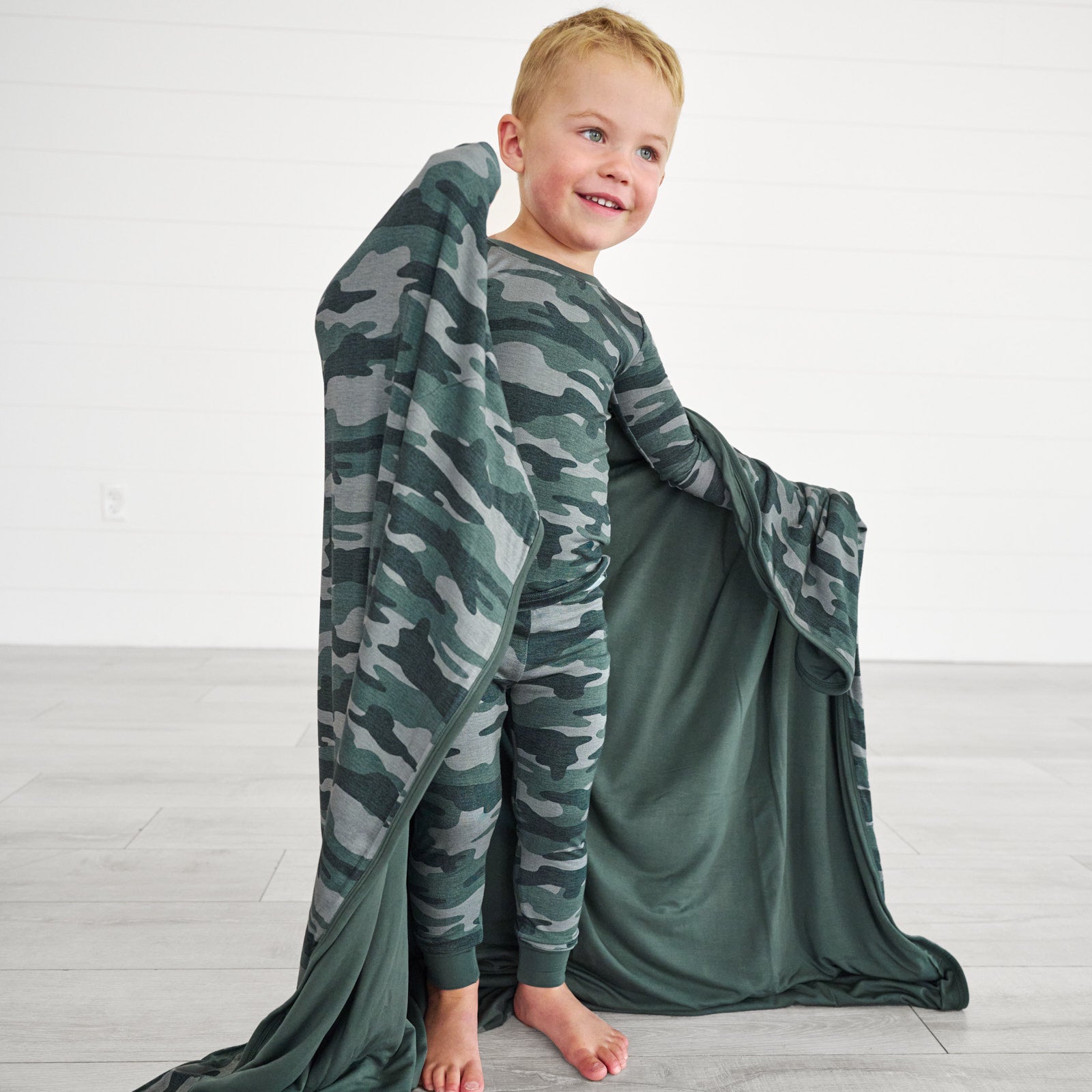 boy posing while displaying the inside detail of the Vintage Camo Large Cloud Blanket® while wearing the Vintage Camo Two-piece Pajama Set