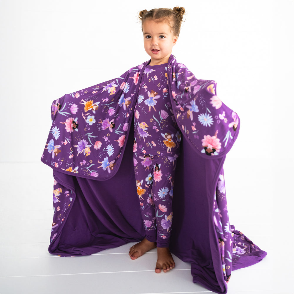 Girl posing while wearing the Garden Fairies Two-piece Pajama Set and wrapped in the Garden Fairies Fitted Crib Sheet, displaying the inside purple color