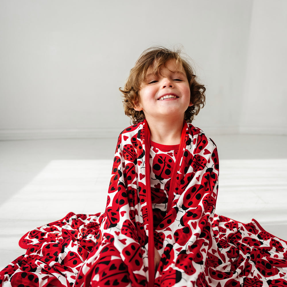 Click to see full screen - Child sitting on the ground wrapped up in a Love Bug printed large cloud blanket