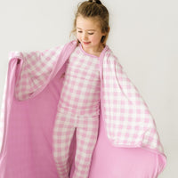 Close up image of a child wearing a Pink Gingham cloud blanket over her shoulders and a matching pink gingham two piece pajama set