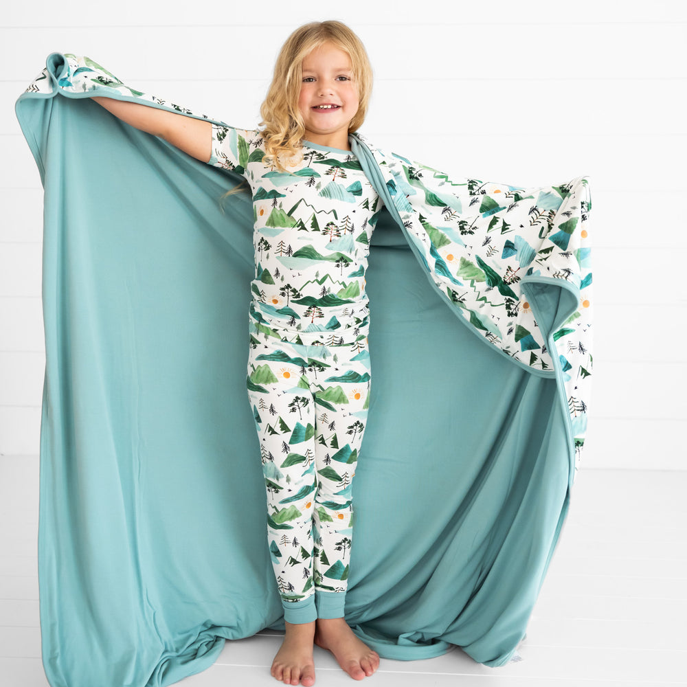 child holding up a Mountain Mist large cloud blanket showing the solid blue backing. She is wearing a matching Mountain Mist two piece short sleeve pj set