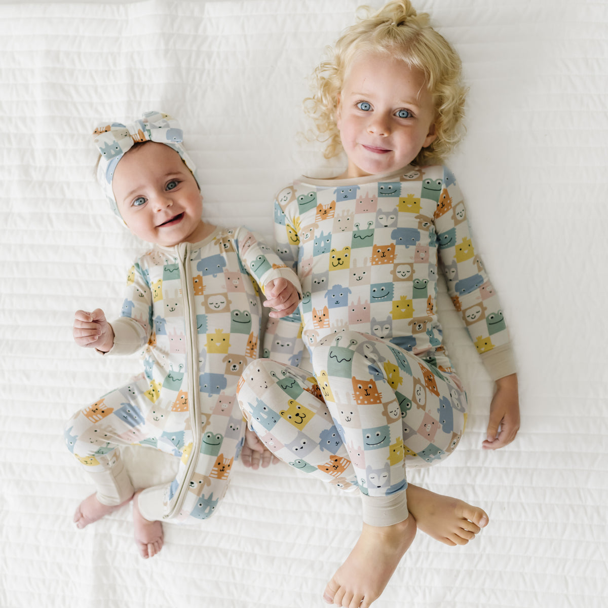 Two children laying on a bed wearing matching Check Mates pjs in zippy and two piece styles paired with a luxe bow headband