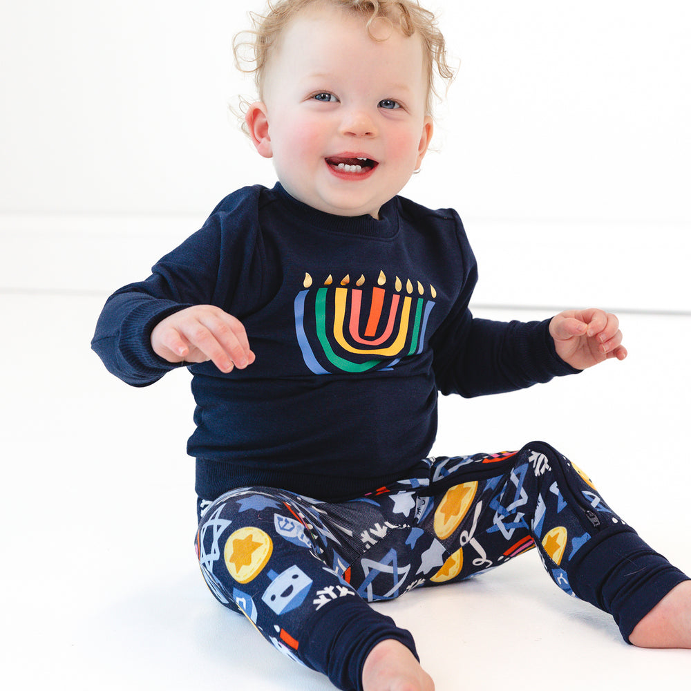 Child sitting on the ground wearing a Menorah Graphic Crewneck over a Playful Dreidels Zippy