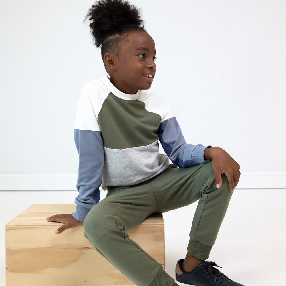 Boy posing while sitting on wooden box and wearing the Olive Panel Crewneck and Olive Jogger