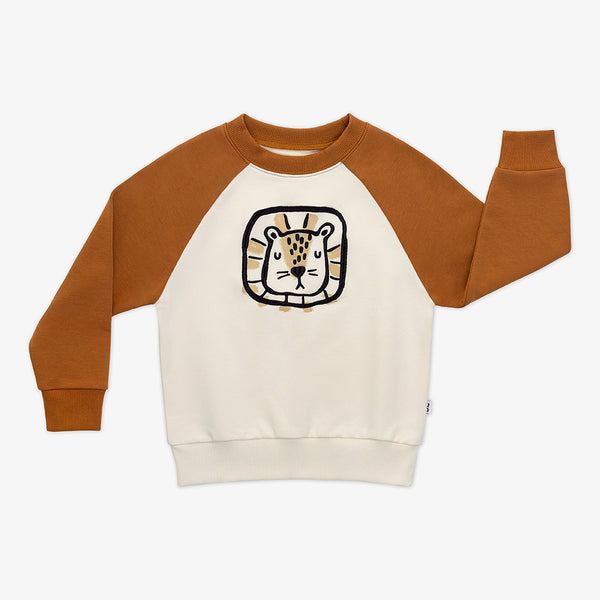 Flat lay of an Arctic raglan crewneck featuring tan sleeves and a black and tan lion illustration on an off-white background