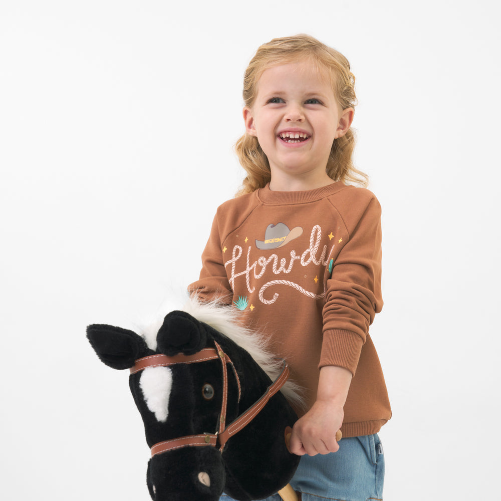 Child wearing a Howdy crewneck sweatshirt holding a toy stick horse