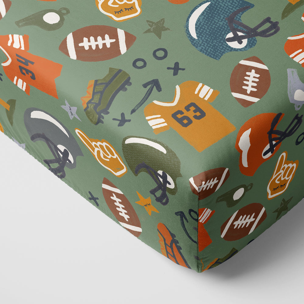 Close-up image of a corner on the crib sheet with a football theme, featuring illustrations of jerseys, helmets, footballs, and other sports-related icons on a green background.