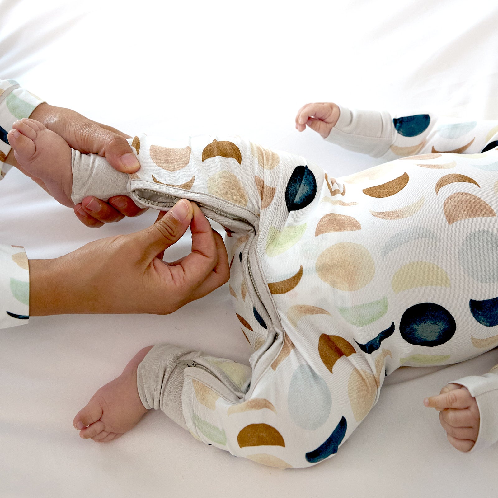 baby laying down wearing the crescent zippy in luna neutral while a parent zips it up