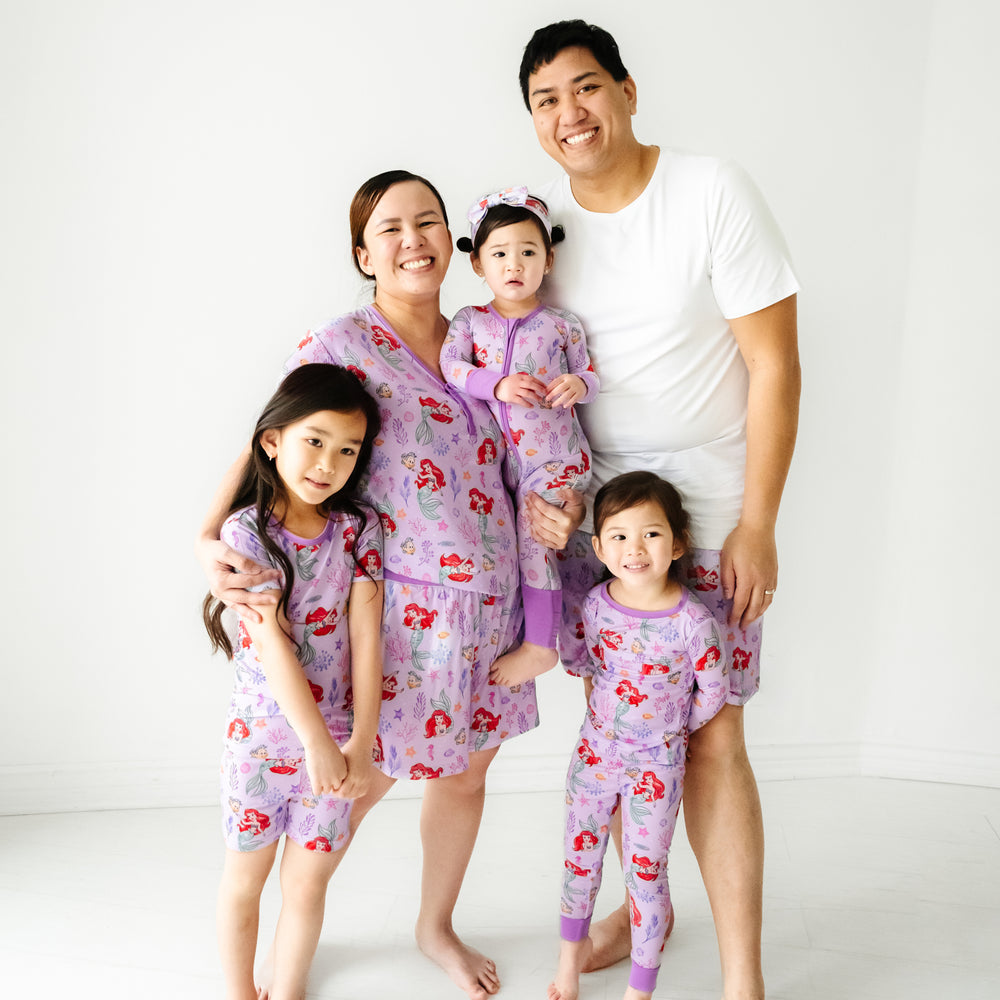 Family of five wearing matching Disney Part of Her World pajama sets. Dad is wearing men's Bright White pajama top paired with Men's Part of Her World pajama shorts. Mom is wearing  Part of Her World women's pajama top and matching women's pajama shorts. Kids are wearing Part of Her World pajamas in two piece and zippy styles