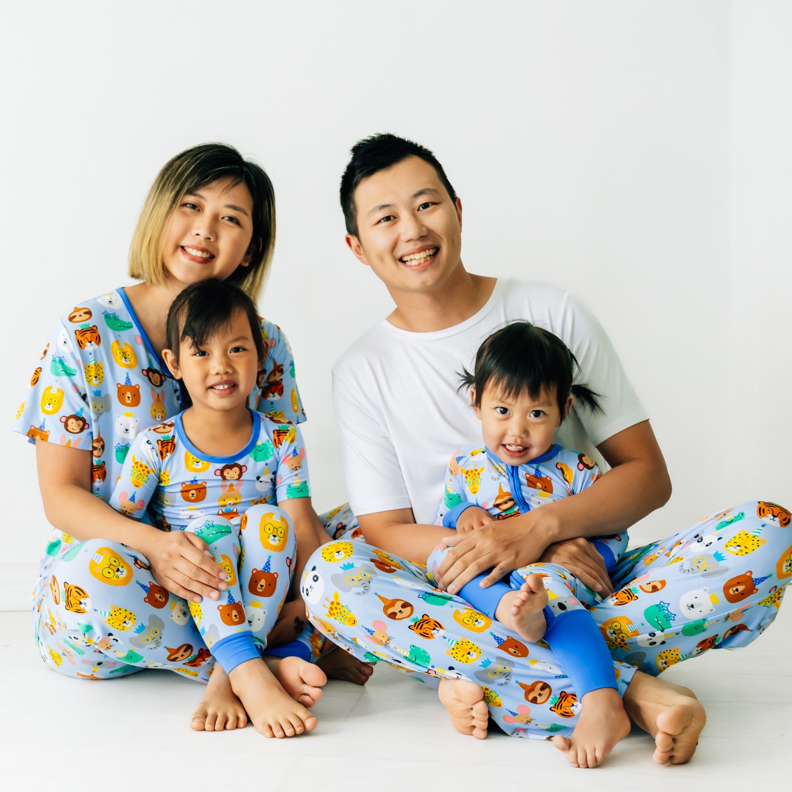 Family of four wearing matching Party Pals pjs. Dad is wearing men's Party Pals men's pj pants paired with a men's bight white top. Mom is wearing Party Pals women's pj top and matching women's pj pants. Kids are wearing Party Pals in two piece and zippy styles.