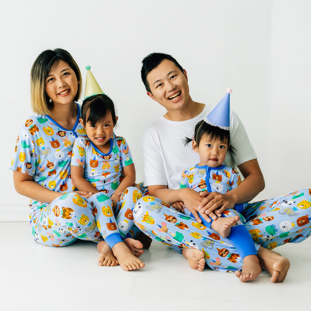 Family of four wearing matching Party Pals pjs. Dad is wearing men's Party Pals men's pj pants paired with a men's bight white top. Mom is wearing Party Pals women's pj top and matching women's pj pants. Kids are wearing Party Pals in two piece and zippy styles.