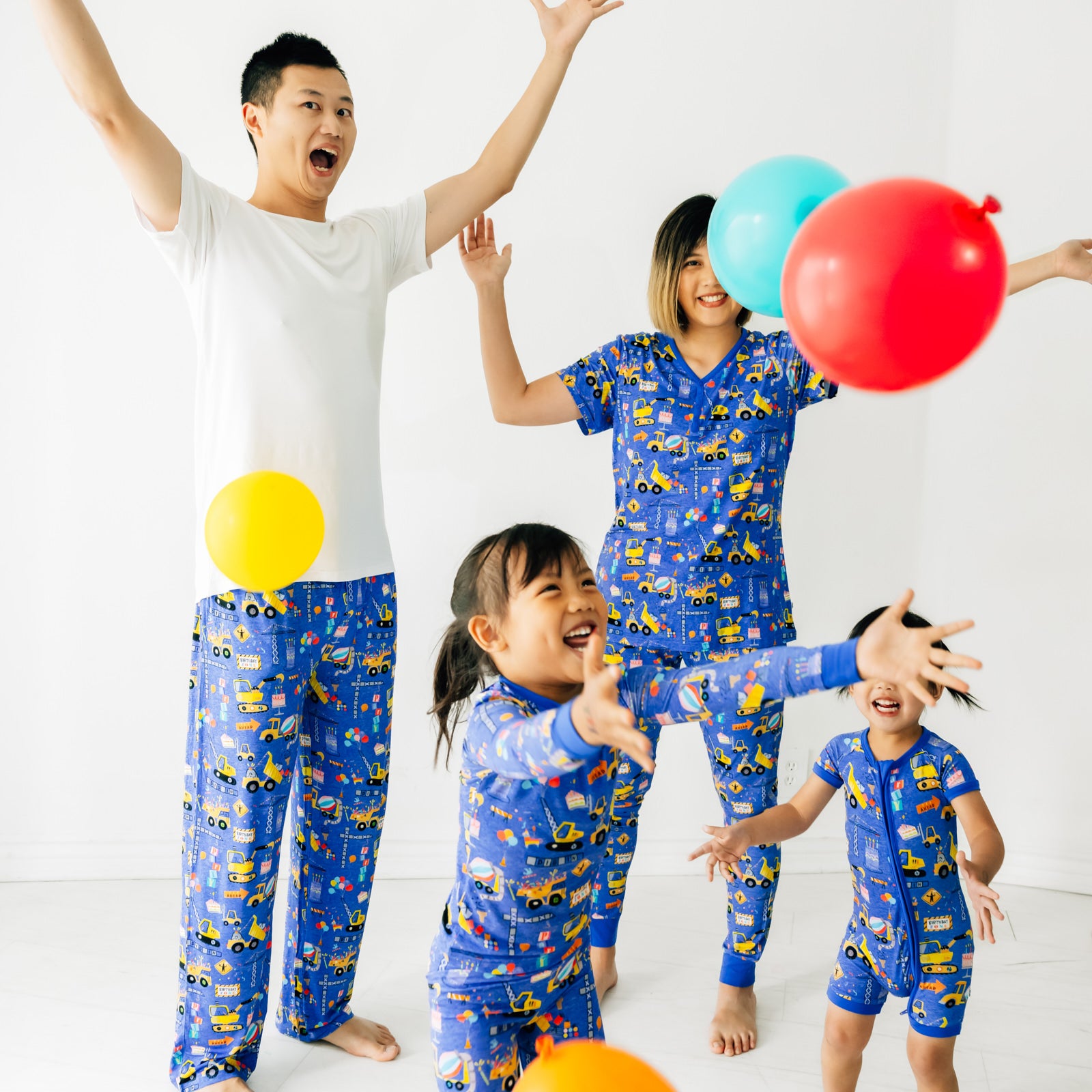Family of four wearing matching Birthday Builder pjs. Dad is wearing men's Birthday Builders men's pj pants paired with a men's bight white top. Mom is wearing Birthday Builders women's pj top and matching women's pj pants. Kids are wearing Birthday Builders in two piece and zippy styles.