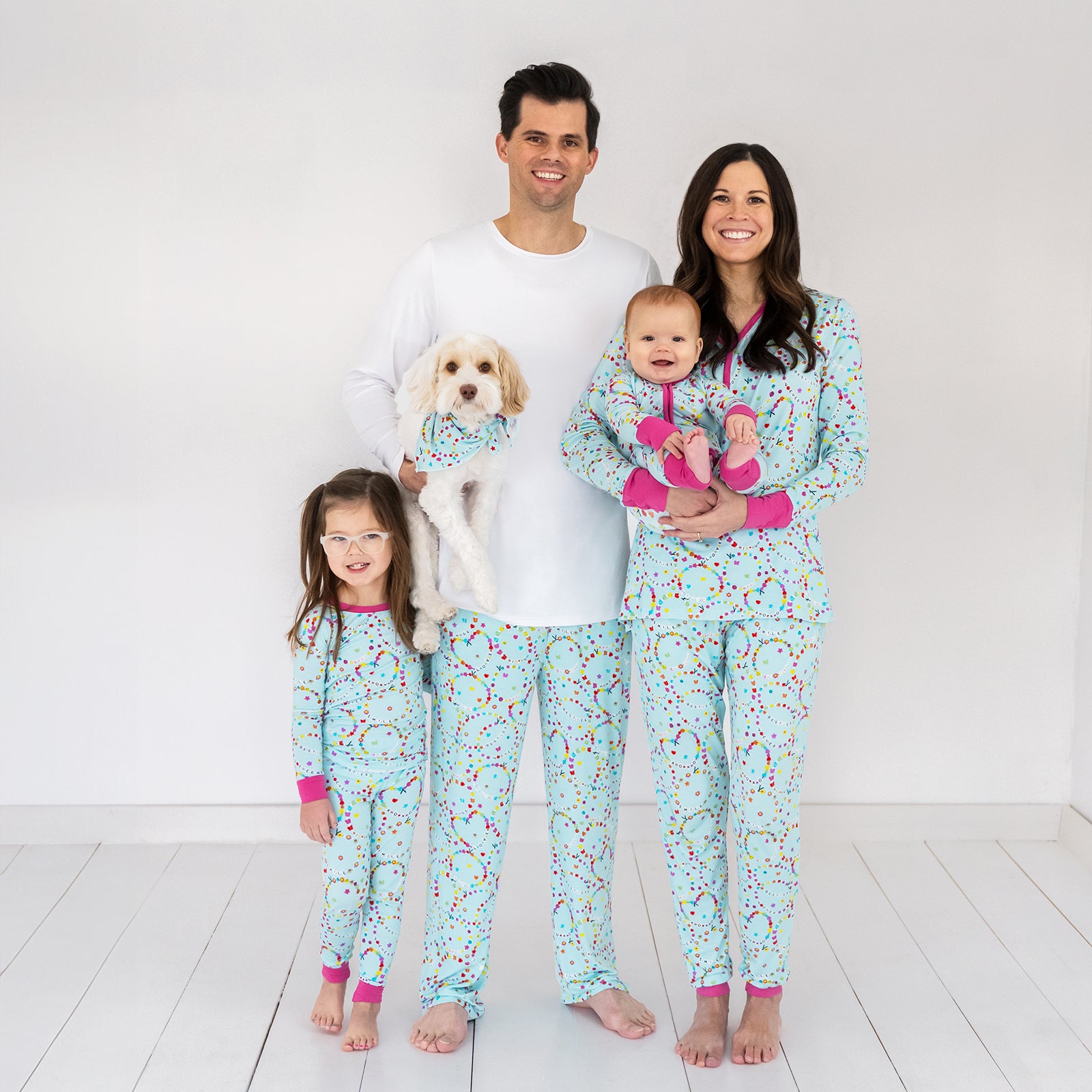 Family of four and their dog wearing matching Friends Forever pjs. Mom is wearing women's Friends Forever pj top and coordinating women's pj pants. Dad is wearing a men's white pj top and men's Friends Forever pj pants. Their children are wearing a Friends Forever zippy and matching two piece pj set. The dog is wearing a matching pet bandana.