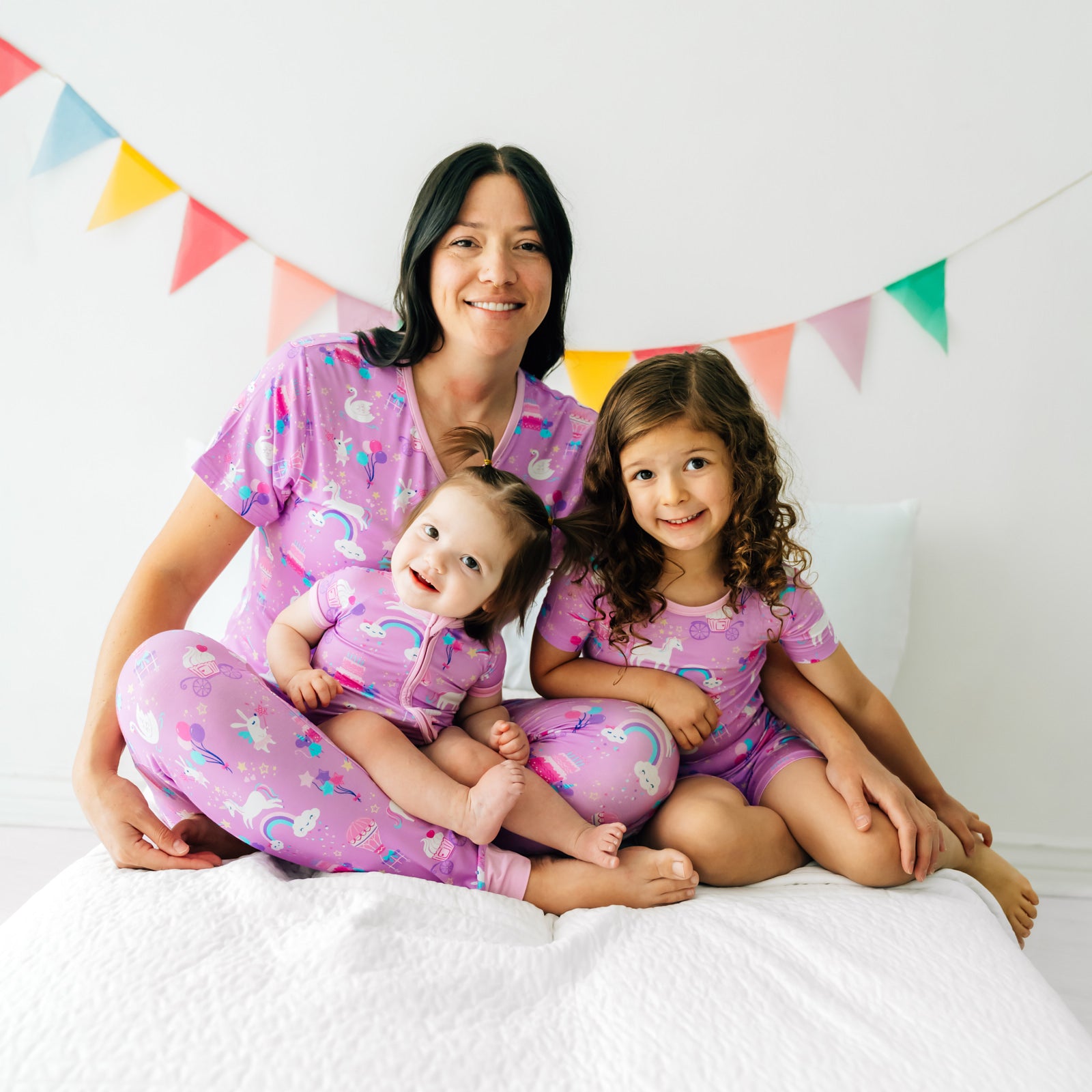 Mother and two daughters sitting together wearing matching Magical Birthday pjs. Mom is wearing women's Magical Birthday pj top and matching pj pants. Children are wearing magical birthday two piece pj set and shorty zippy